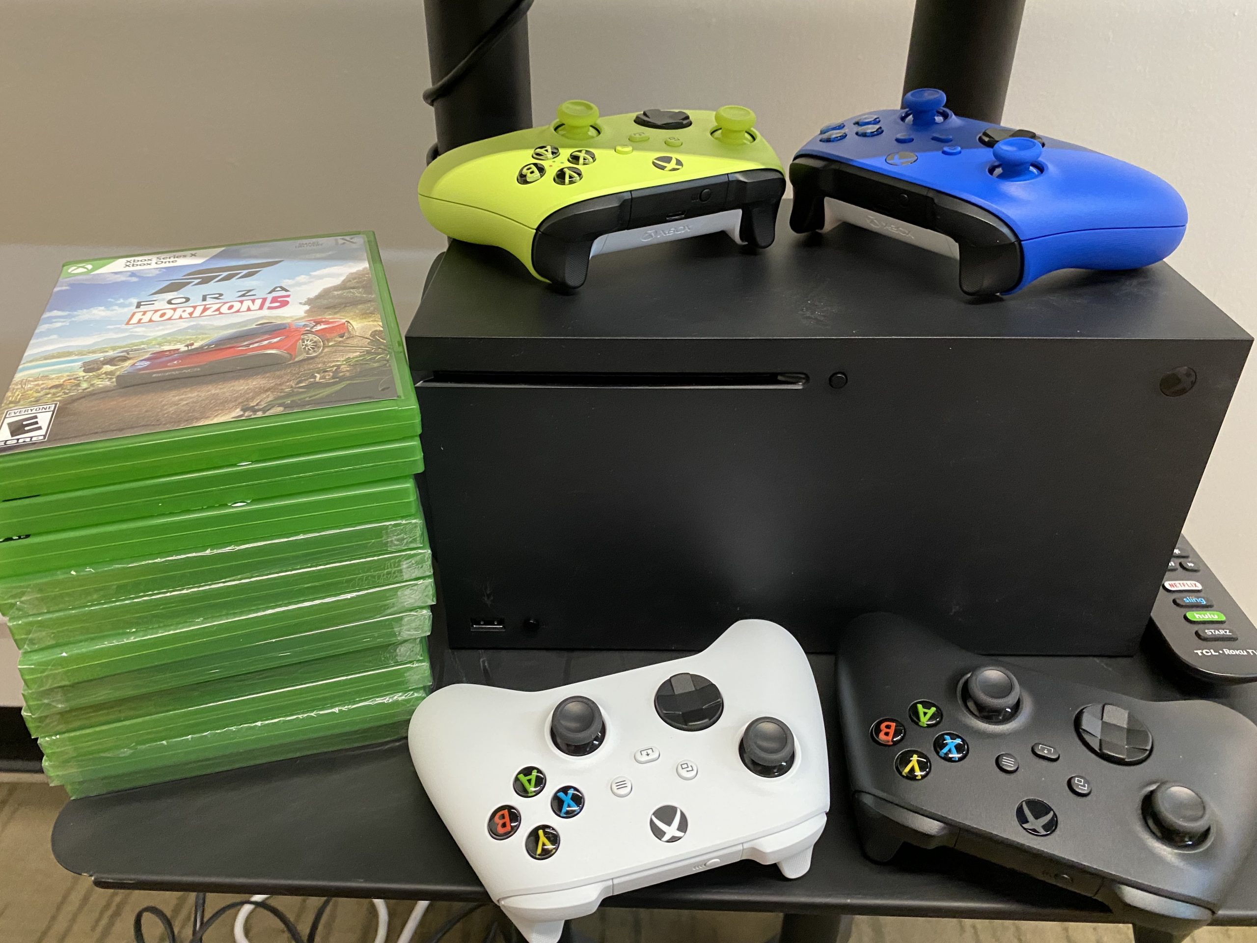Xbox series X gaming system with black, whitem blue, and neon green controllers next to a stack of games