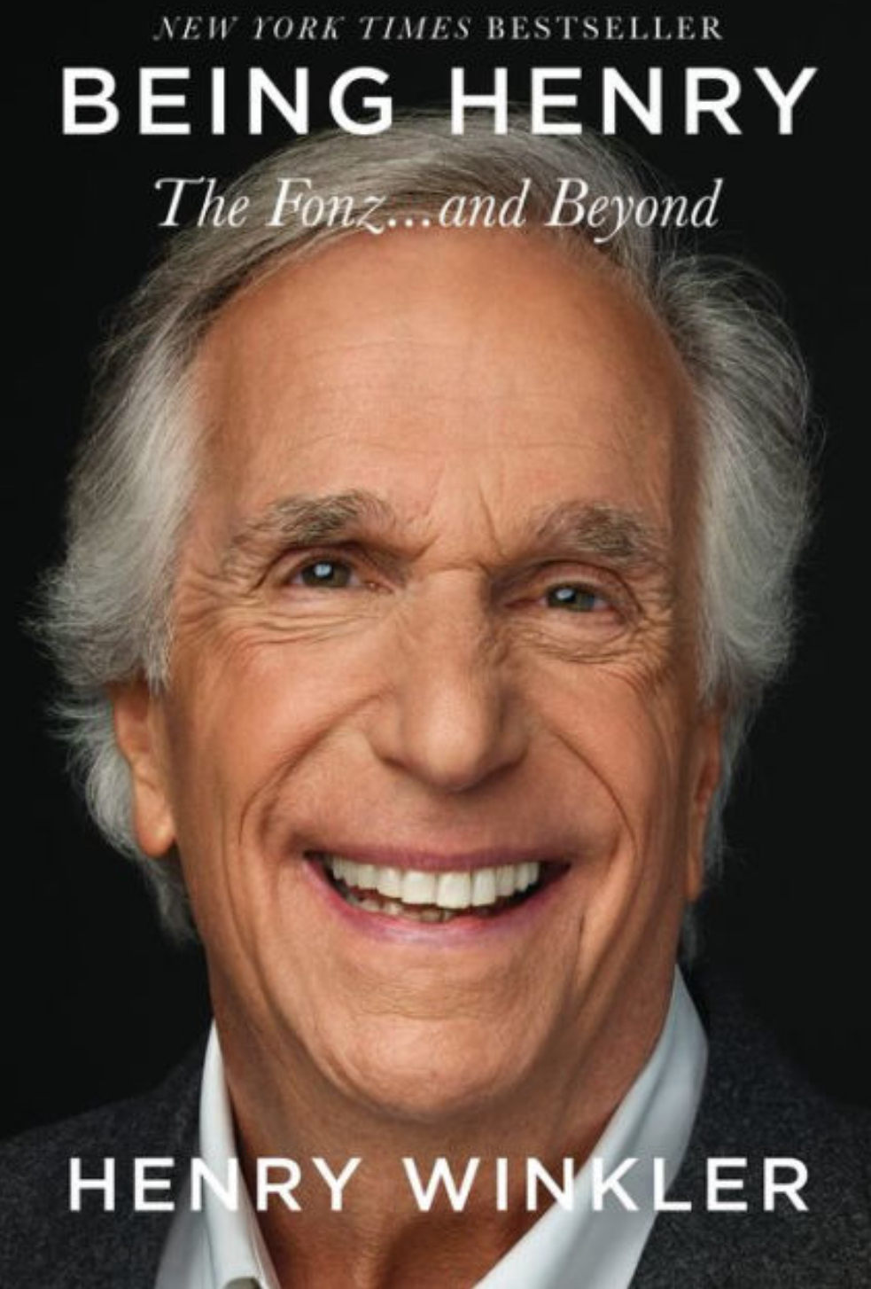 Book cover prominently showing recent photo of actor Henry Winkler smiling with grey hair and wearing an unbuttoned white dress shirt with a dark suit jacket. Behind Henry is a dark background and these words are on the book cover: New York Times Bestseller (centered at the top in white letters.) Below are these words in large white all capital letters: BEING HENRY Below in white italics are these words: The Fonz…and Beyond Centered at the bottom in slightly smaller white all capital letters is the name, HENRY WINKLER